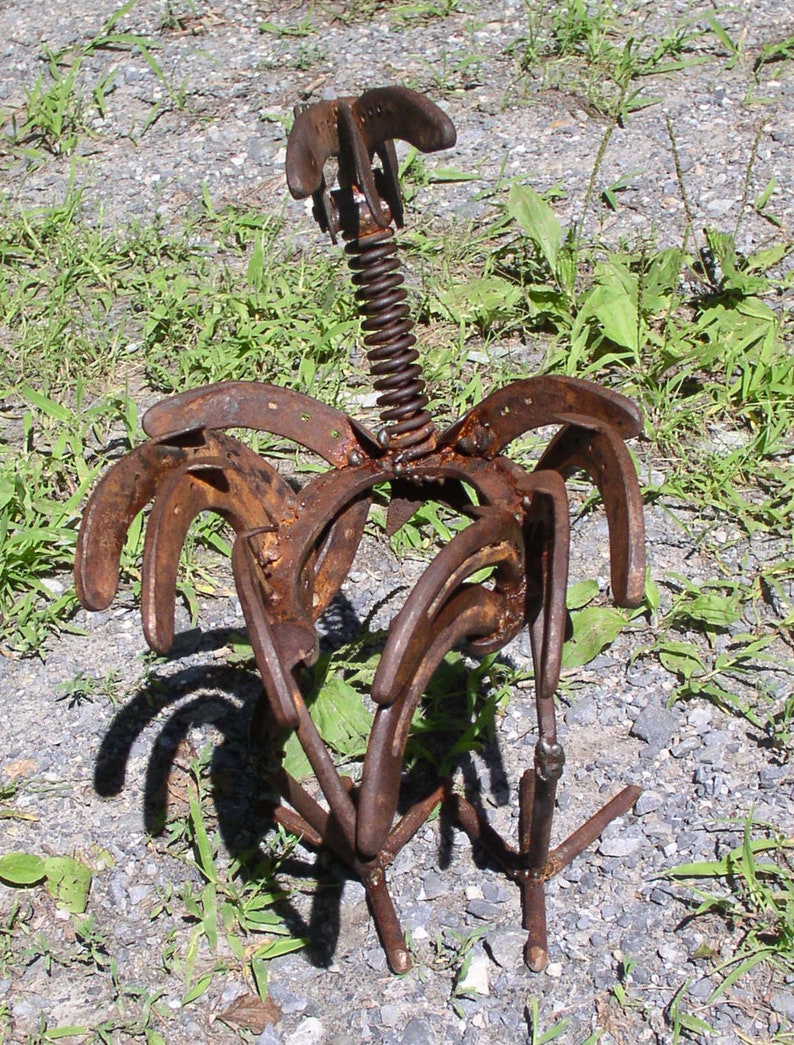 K&ouml;nnte beinhalten: Eine rostige Metallskulptur eines Vogels, gefertigt aus Hufeisen und einer Feder. Die Skulptur hat eine verwitterte, braune Oberfl&auml;che und steht auf Kies und Gras. Der Vogelk&ouml;rper wird durch gebogene Hufeisen geformt.
