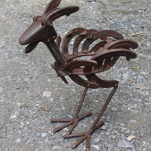 K&ouml;nnte beinhalten: Rostige Metallskulptur eines Vogels, hergestellt aus Hufeisen und anderen Metallteilen. Der Vogel hat einen langen Hals mit einer Feder, einen Schnabel und einen Schwanz aus gebogenen Hufeisen. Die Skulptur ist braun und steht auf zwei Metallbeinen.