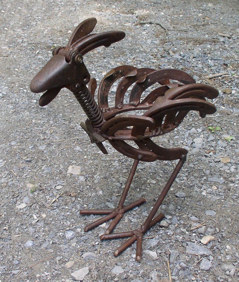 K&ouml;nnte beinhalten: Rostige Metallskulptur eines Vogels, hergestellt aus Hufeisen und anderen Metallteilen. Der Vogel hat einen langen Hals mit einer Feder, einen Schnabel und einen Schwanz aus gebogenen Hufeisen. Die Skulptur ist braun und steht auf zwei Metallbeinen.