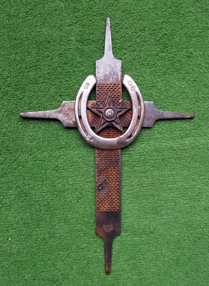 Puede incluir: Una cruz de metal oxidado hecha de una lima y una herradura. La herradura tiene una estrella en el centro y las palabras "5/8" y "0" en los lados. La lima tiene las palabras "Barbed Steel" y "USA" en ella.