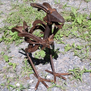 K&ouml;nnte beinhalten: Rostige Metall-Hahn-Skulptur aus recycelten Hufeisen und Metallteilen gefertigt. Die Skulptur hat einen Federk&ouml;rper, ein sternf&ouml;rmiges Brustdetail und ein verwittertes, rostiges Finish. Der Hahn steht auf einem Kiesboden mit gr&uuml;nen Grasb&uuml;scheln.
