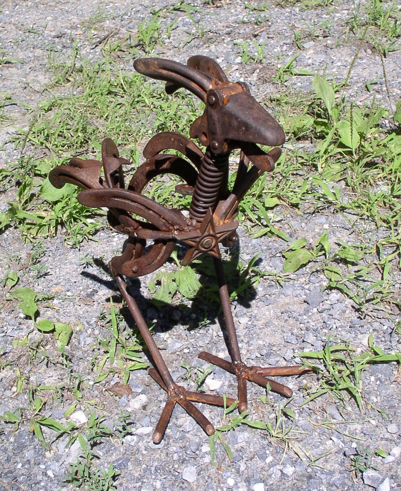 K&ouml;nnte beinhalten: Rostige Metall-Hahn-Skulptur aus recycelten Hufeisen und Metallteilen gefertigt. Die Skulptur hat einen Federk&ouml;rper, ein sternf&ouml;rmiges Brustdetail und ein verwittertes, rostiges Finish. Der Hahn steht auf einem Kiesboden mit gr&uuml;nen Grasb&uuml;scheln.