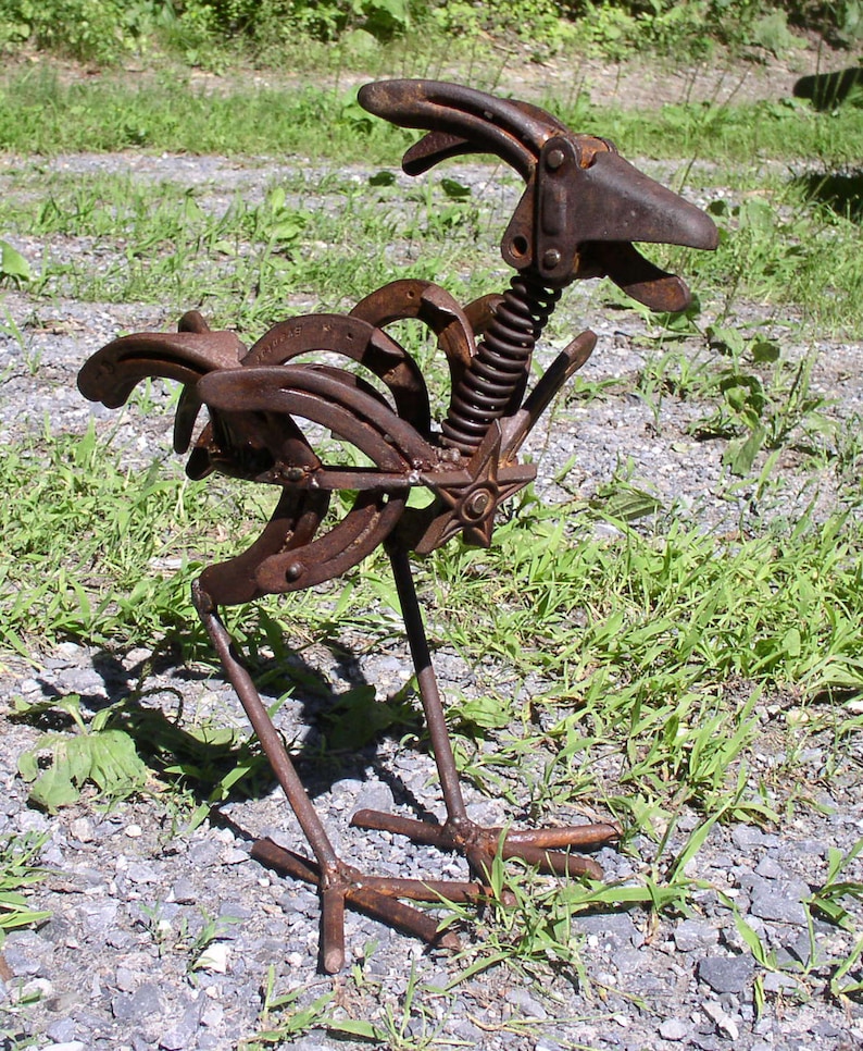 K&ouml;nnte beinhalten: Eine rostige Metallskulptur eines Vogels, hergestellt aus Hufeisen und anderen Metallteilen. Die Skulptur hat eine Feder f&uuml;r den Hals und ein sternf&ouml;rmiges Element. Der Vogel steht auf einer Schotteroberfl&auml;che mit Gras im Hintergrund.