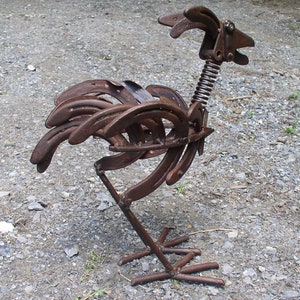 K&ouml;nnte beinhalten: Rostige Metall-Hahn-Skulptur aus Hufeisen gefertigt. Die Skulptur hat einen Federhals, einen detaillierten Kopf und einen K&ouml;rper aus gebogenen Hufeisen. Der Hahn steht auf einem Metallsockel und zeigt ein einzigartiges, handgefertigtes Design.