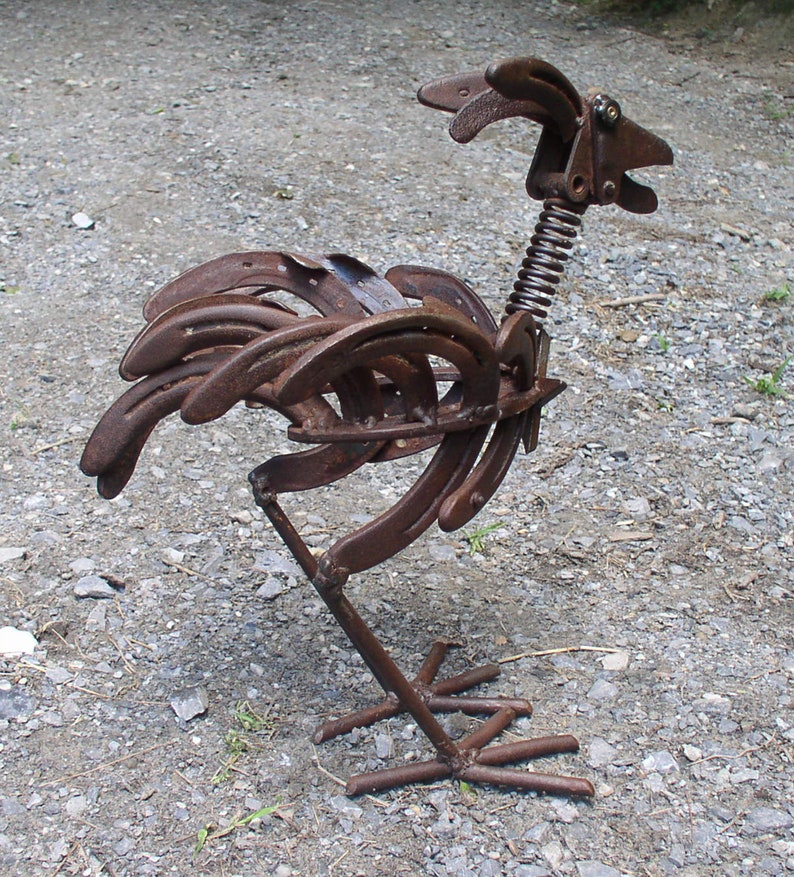K&ouml;nnte beinhalten: Rostige Metall-Hahn-Skulptur aus Hufeisen gefertigt. Die Skulptur hat einen Federhals, einen detaillierten Kopf und einen K&ouml;rper aus gebogenen Hufeisen. Der Hahn steht auf einem Metallsockel und zeigt ein einzigartiges, handgefertigtes Design.