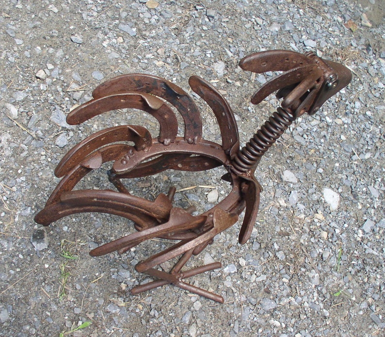 K&ouml;nnte beinhalten: Rustikale Metall-Hahn-Skulptur aus Hufeisen gefertigt. Die Skulptur hat eine Feder f&uuml;r den Hals und steht auf einer Kiesoberfl&auml;che. Der K&ouml;rper und der Schwanz des Hahns bestehen aus gebogenem Metall, das ein einzigartiges, handgefertigtes Design zeigt.