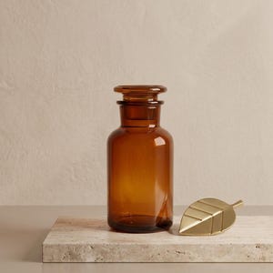 Peut inclure: Une bouteille d'apothicaire en verre brun avec un bouchon, à côté d'un objet en forme de feuille de couleur or. La bouteille est cylindrique avec une base arrondie. La feuille a des nervures détaillées. Les objets sont sur une surface en pierre claire.