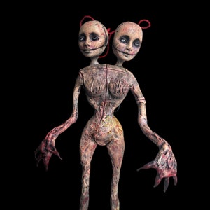 May include: A disturbing sculpture of a conjoined figure with two heads. The heads have pale skin, black stitched smiles, and red string tied to the top. The body is a mix of pink, brown, and red, with a textured appearance and outstretched arms.