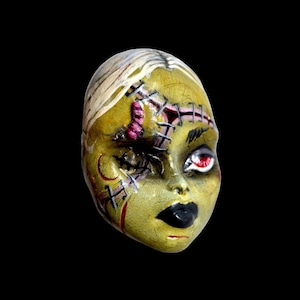 May include: A hand-painted doll head with a stitched-up face, one eye is closed and the other is red. The doll has a green face with pink and red stitching and a black mouth.