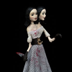 May include: A handcrafted doll with two heads, dressed in a white off-the-shoulder top, a black and white striped skirt, and a black corset. The doll holds a bloody knife and wears black gloves. It has black hair and pale skin.