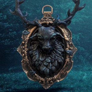 Puede incluir: Una escultura de cabeza de ciervo negra con cuernos, enmarcada en un marco dorado adornado, con una textura de hojas azules y verdes. La escultura está colgada sobre un fondo azul oscuro.