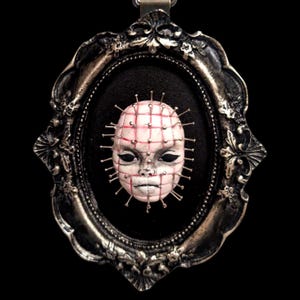 May include: A framed miniature sculpture of a doll's head with a grid pattern and many pins sticking out. The doll's head is painted pink and white with black eyes.