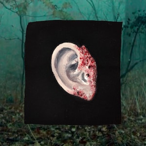 May include: A painting of a human ear on a black canvas. The ear is rendered in shades of white and gray, with a section of the ear detailed with red and white paint. The background is a blurred forest scene.