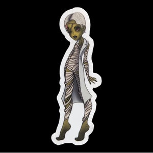 May include: A sticker featuring a cartoon illustration of a green-skinned, white-haired mummy wearing a white lab coat and bandages. The mummy has a stitched-up scar on its forehead and is standing with its arms slightly raised.