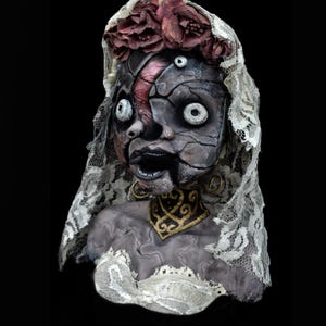 May include: A haunting doll bust with a cracked face, adorned with a lace veil and a floral crown. The doll's features include wide, unsettling eyes and a dark, open mouth. A gold and black collar adds a touch of elegance to the eerie design.