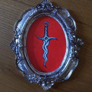 May include: An oval-shaped, ornate silver-toned frame with intricate floral detailing surrounds a vibrant red background. Centered on the red field is a detailed illustration of a wavy-bladed dagger with a textured handle and a circular pommel.