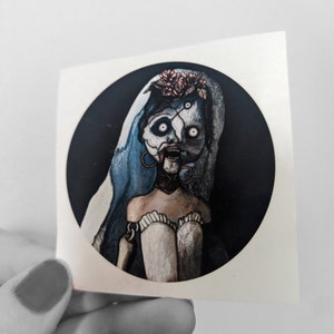 May include: A round sticker featuring a bride doll with a blue veil, a white dress, and a floral crown. The doll has a pale face with dark eyes and a wide open mouth. The background is black.