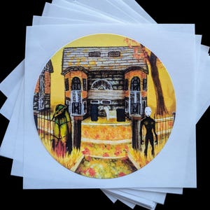 May include: A circular sticker with a hand-painted illustration of a house with a yellow, orange, and brown color scheme. The house has a gray roof and two windows on each side. Two figures stand in front of the house, one in green and one in black. The background is a yellow and orange color with a tree on the right side.