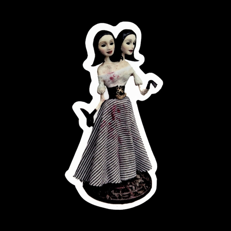 May include: A two-headed doll with black hair and white and black striped dress. The doll is wearing a black belt with a gold buckle. The doll is standing on a black and white base.