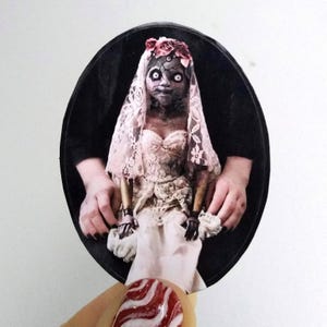 May include: A porcelain doll wearing a white lace wedding dress and a floral crown. The doll has a pale complexion and dark eyes. The doll is sitting in a chair with its hands resting on its lap.