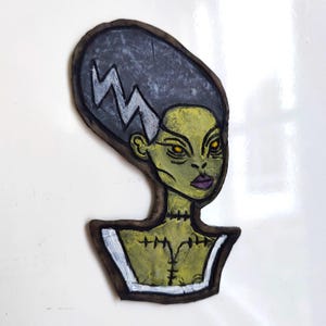 May include: Hand-painted wooden cutout of a green-skinned character with dark gray hair and a white lightning bolt. The character has yellow eyes and purple lips. The cutout has a brown border and a white collar.