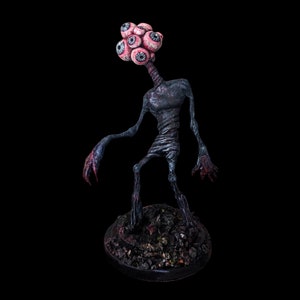 May include: A dark, textured sculpture of a creature with multiple eyes on its head, a long neck, and thin limbs. The figure stands on a dark, textured base against a black background. The creature's hands and feet have a reddish hue.