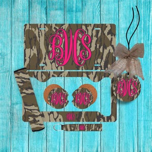 May include: A camouflage-patterned car accessory set featuring a license plate, steering wheel cover, coasters, key chain, and a car air freshener. Each item has a hot pink monogram. The set is displayed on a turquoise wooden surface.