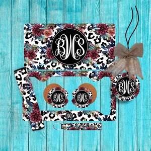 May include: A set of car accessories featuring a white and black leopard print with burgundy floral accents. The collection includes a license plate, a license plate frame, two circular cup coasters, a keychain, and a round ornament with a burlap bow.