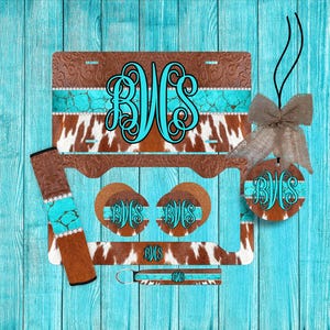 May include: A set of vehicle accessories featuring a brown leather-look pattern, white cowhide print, and a turquoise stone design. The items include a license plate frame, two circular coasters, a keychain, and a strap, all displaying the monogram BVS.