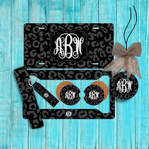 May include: A set of black and white leopard print car accessories with the monogram "ABW". The set includes a license plate holder, two coasters, a key chain, and a car air freshener.