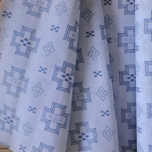 Pode incluir: Tecido branco com um padrão geométrico azul repetitivo. O padrão apresenta formas de cruz e desenhos menores em forma de diamante. O tecido é drapeado, mostrando dobras e textura, com um fundo de madeira.