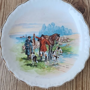 Puede incluir: Un plato de cerámica blanco con borde dorado presenta una escena pintada a mano de un cazador con un perro y un caballo en un entorno de bosque. La escena está rodeada de un cielo azul y blanco con nubes.