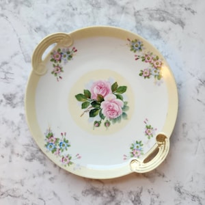 May include: A white porcelain serving plate with a yellow rim and two gold handles. The plate features a hand-painted floral design with pink roses and blue flowers.
