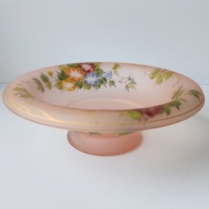 Puede incluir: Un cuenco de pedestal de vidrio rosa con un diseño floral. El cuenco tiene un borde dorado y un delicado patrón floral pintado en el lateral.
