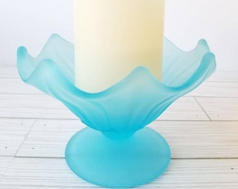 May include: A frosted blue glass candle holder with a scalloped edge and a round base. A white pillar candle is sitting in the holder.