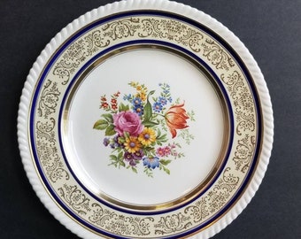 May include: A white porcelain plate with a floral design. The plate features a gold rim, a blue border, and a white rope-like edge. The center of the plate displays a bouquet of colorful flowers, including pink roses and orange tulips.