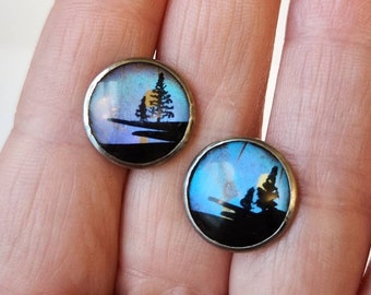 May include: A pair of round earrings featuring a blue and purple iridescent butterfly wing design. The earrings depict a silhouette of a tree and a sun against a black background.
