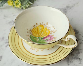 May include: A yellow teacup and saucer set with a floral design featuring a yellow rose and a pink rose. The saucer has a yellow band with gold trim.