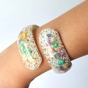 May include: A white resin bangle bracelet with a glittery finish and colorful embedded pieces. The bracelet is open and has a curved shape.