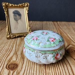 May include: A small, white porcelain trinket box with a floral design featuring pink roses and green accents. The box has a rounded shape and is decorated with gold trim. A vintage gold-framed portrait is in the background.
