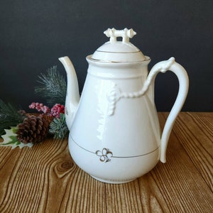 Könnte beinhalten: Eine weiße Porzellan-Teekanne mit Deckel, Ausguss und Henkel. Die Teekanne hat goldfarbene Verzierungen und ein dekoratives Blumendesign. Der Griff hat eine einzigartige geschwungene Form. Die Teekanne steht auf einer Holzoberfläche.