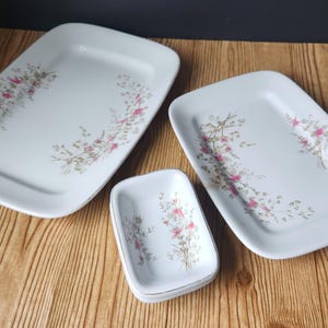 May include: Three white rectangular ceramic serving dishes of varying sizes, each featuring a delicate floral pattern with small pink blossoms and green stems arranged in a curved design along the edges of the smooth, glossy surfaces.