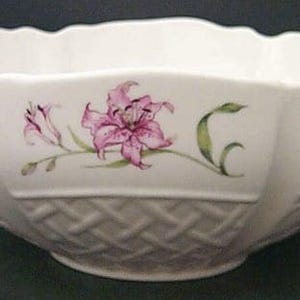 May include: A white ceramic bowl with a scalloped rim and a basket-weave texture around the base. The exterior features a painted floral pattern of pink lilies with green stems and leaves spaced evenly around the circumference of the vessel.