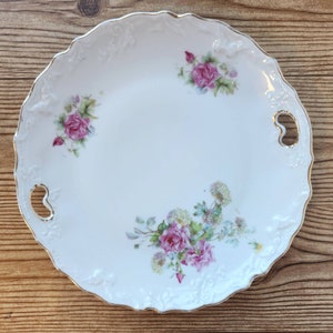 Puede incluir: Un plato de servir de porcelana blanca con un borde dorado y un diseño floral. El plato tiene dos asas a los lados y presenta rosas rosas y verdes.