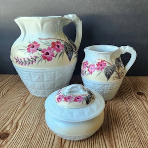 May include: A set of three white ceramic pieces with floral designs. The set includes a pitcher, a creamer, and a sugar bowl with a lid. The floral design features pink flowers and green leaves on a cream background.