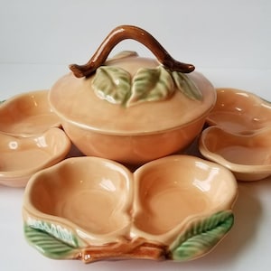 Könnte beinhalten: Eine Servierplatte aus Keramik in Pfirsichfarbe mit Deckel und vier kleineren Schalen. Die größere Schale hat einen braunen Zweiggriff und grüne Blätter. Die kleineren Schalen sind pfirsichförmig und haben grüne Blätter.