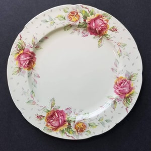 May include: A white ceramic plate with a scalloped edge, adorned with a floral rose pattern. The roses are pink and yellow, surrounded by green leaves and a light gray dotted background. The plate is a classic piece of tableware.