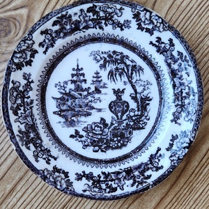 May include: A white ceramic plate with a black and blue floral design. The centre of the plate features a black and white illustration of a pagoda and a tree.