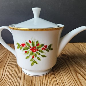 May include: White porcelain teapot with a lid, handle, and spout. The teapot is decorated with a gold rim and a holly and berry design. The teapot is on a wooden surface.