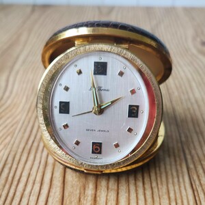 May include: A vintage travel clock with a gold-tone case and a light-colored face. The clock face features black numerals, gold hands, and the text "Seven Jewels." The clock is partially open, revealing a brown leather-like exterior.